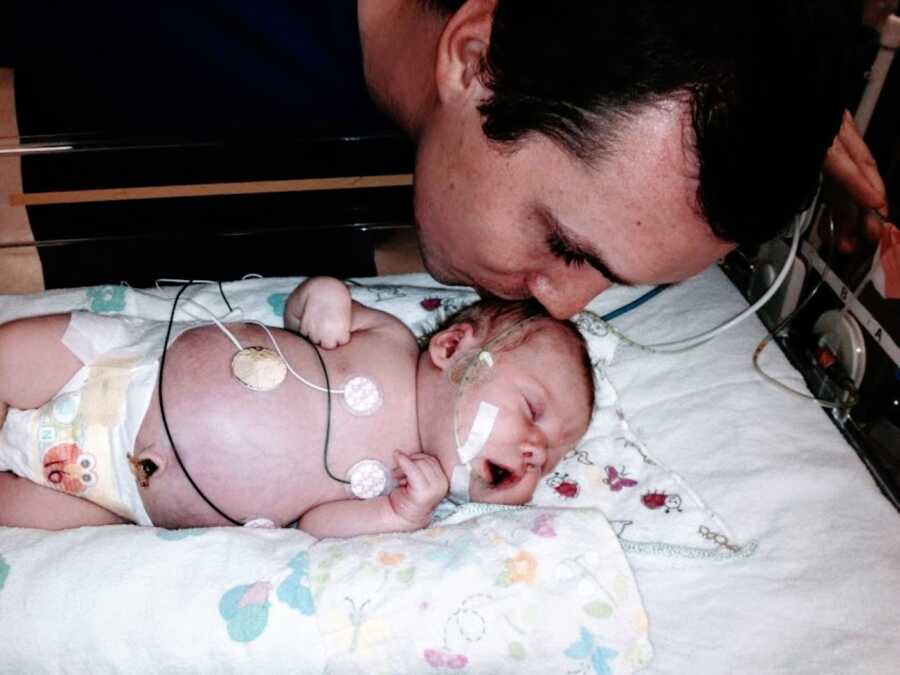 Young new dad kisses his newborn daughter on the head while she's in the NICU