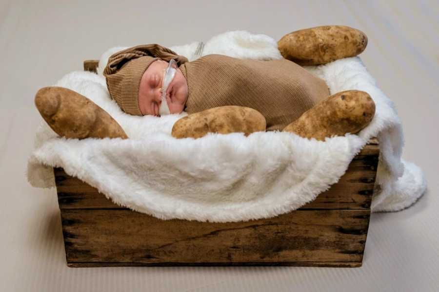 Mom takes newborn photos of firstborn son swaddled in a brown blanket with potatoes surrounding him in a basket