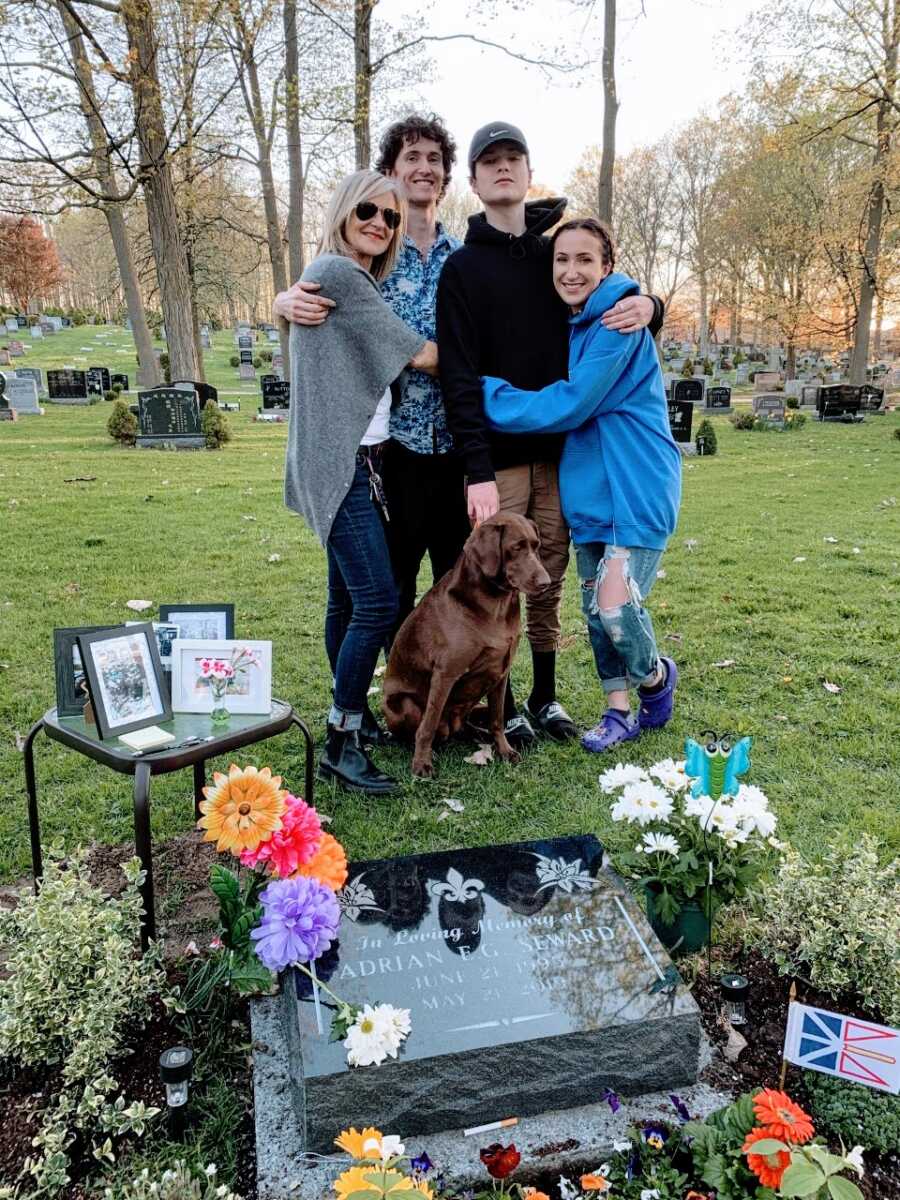 Mom hugs her three living children while standing in front of her oldest son's gravesite