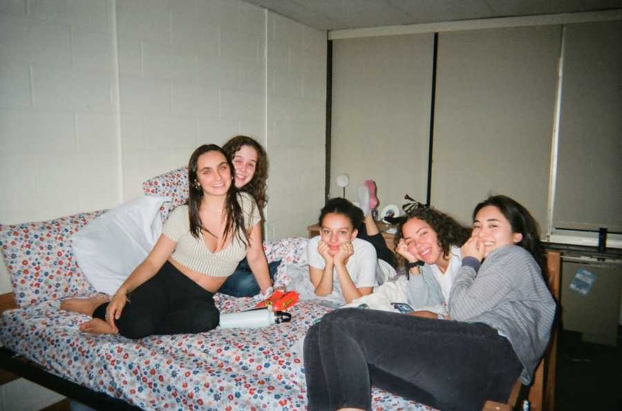 Five girls sit on a bed in a small room