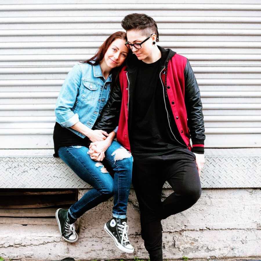 Queer couple take a photo together while holding hands, both wearing jackets and jeans