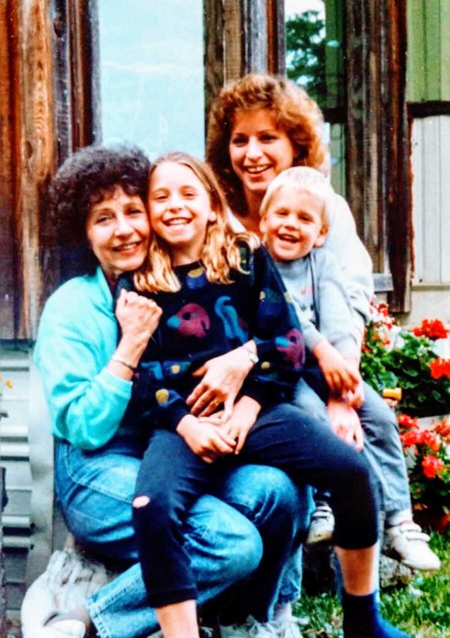 Little girl in an 80's sweater smiles big for a family photo with her little brother, mom, and grandma