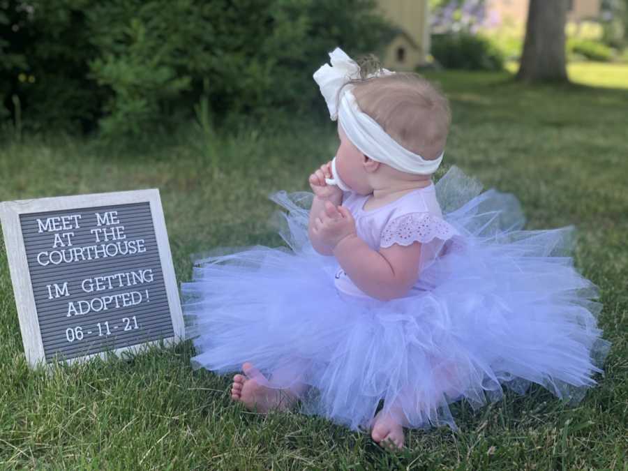 Little girl wears a headband and a tutu while looking at a sign that reads "Meet me at the courthouse, I'm getting adopted!"