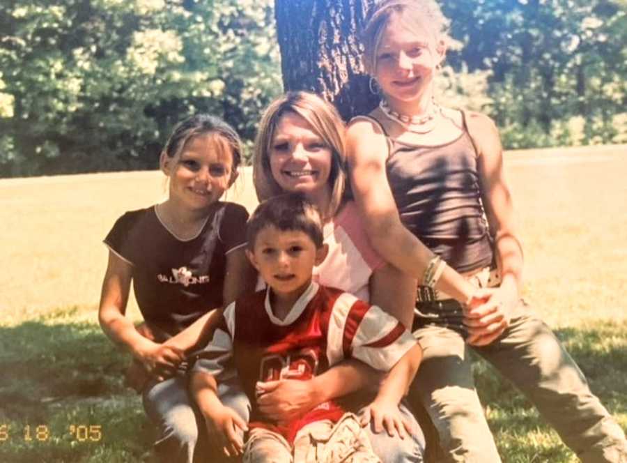 Young single mom of three takes a photo with her kids while they lean against a tree