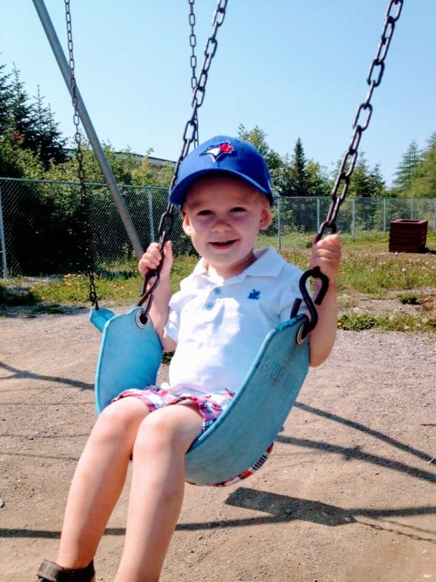 Young little boy in a white polo and blue hat smiles while swinging in his backyard