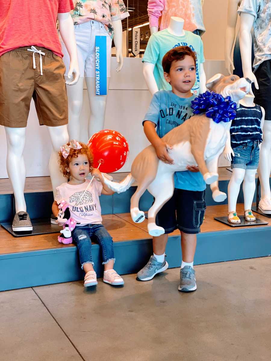 Little boy with autism holds his new Magic dog mannequin from Old Navy while his sister sits in the background holding a red balloon