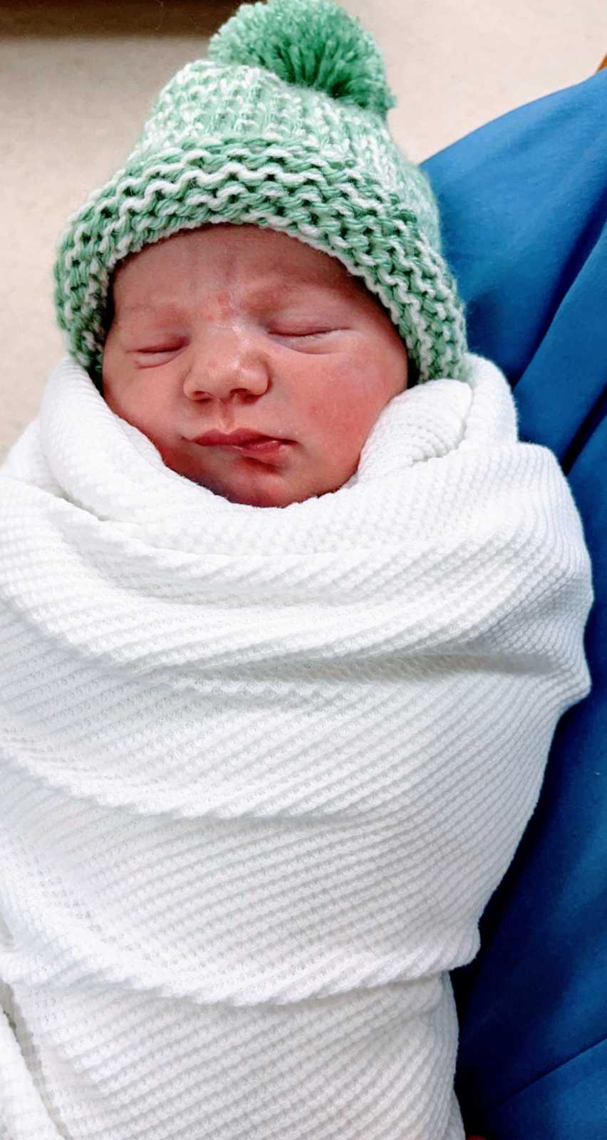 Mom takes photo of her newborn baby swaddled in a white blanket and wearing a green and white knit beanie