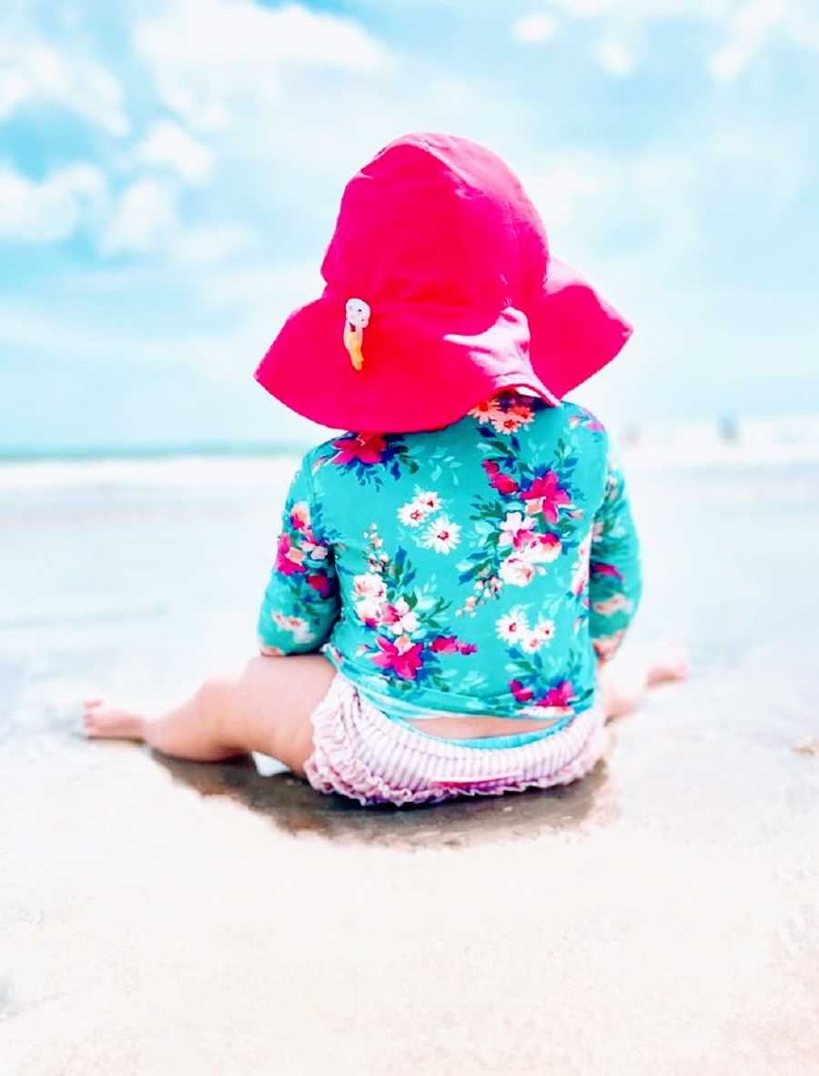 A little girl in a diaper and a sunhat looks out at the water