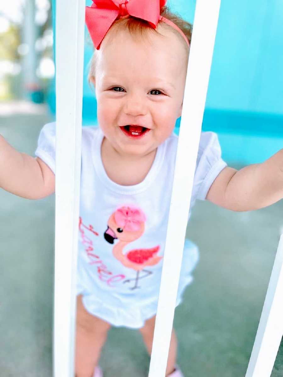 A little girl looks between bars and smiles with two teeth