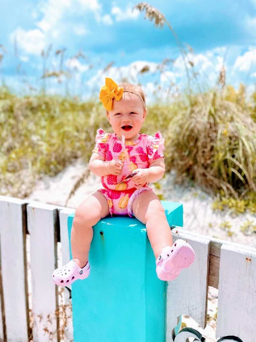 A baby girl sits on a fencepost by the sea