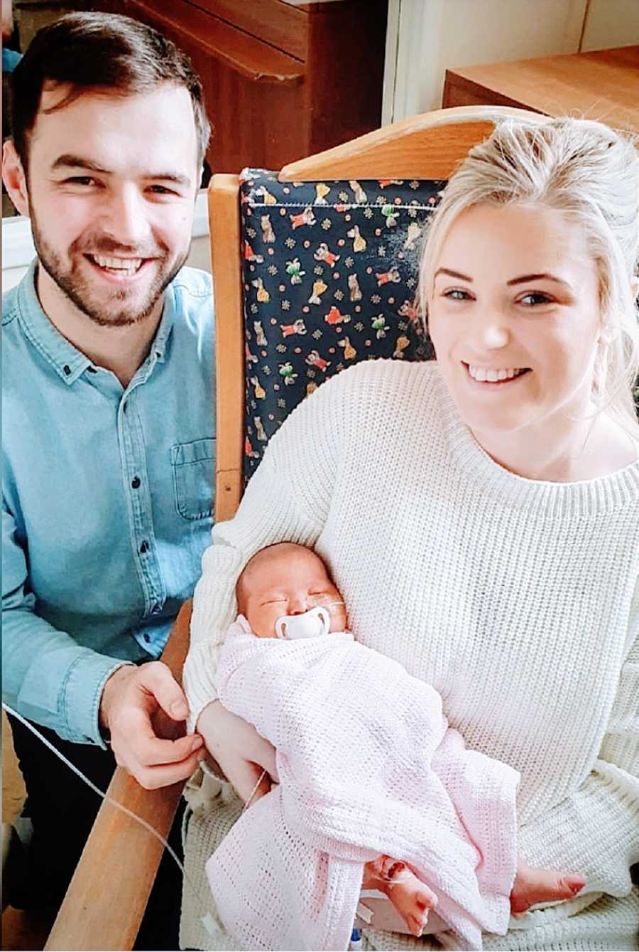 A woman holds her newborn while her partner stands behind