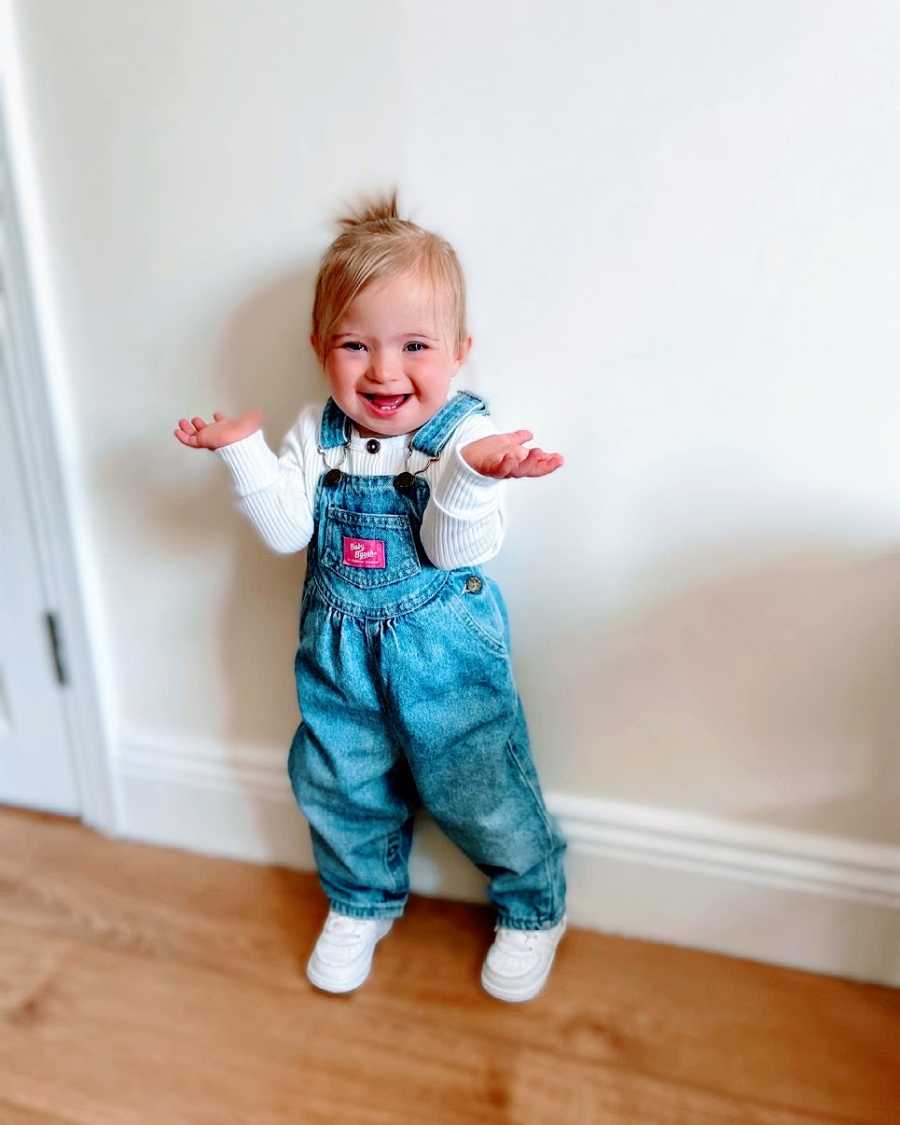 A little girl wearing overalls shrugs her shoudlers