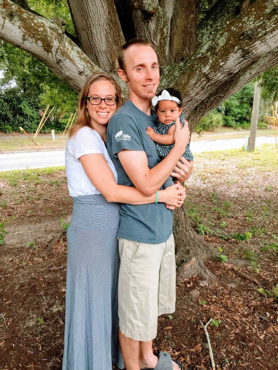 Parents stand together holding their baby girl by a tree