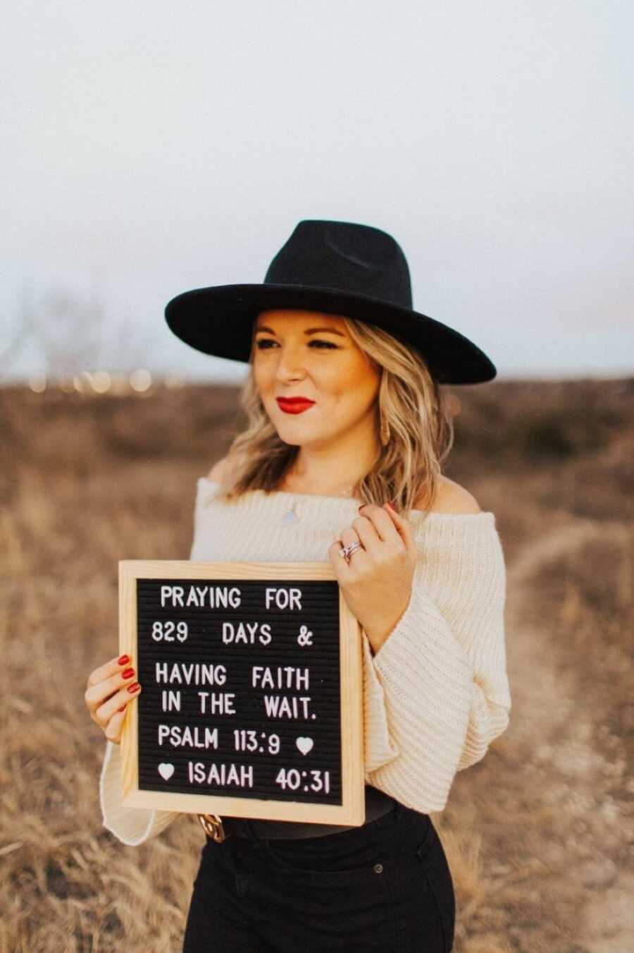 Woman battling unexplained infertility takes a photo in a white sweater and black hat with a sign that says "Praying for 829 days & having faith in the wait"