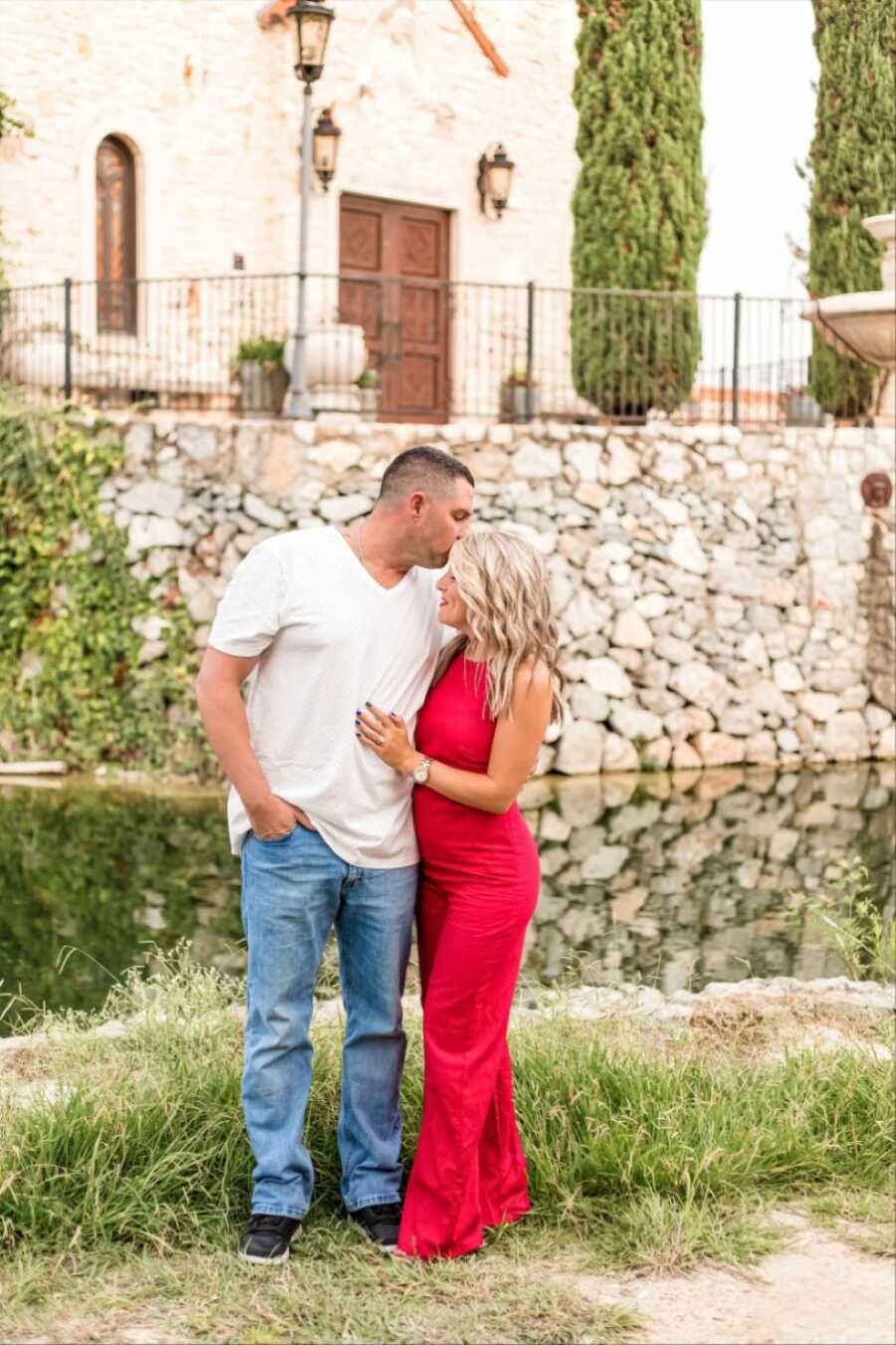 Man wearing a white t-shirt and blue jeans kisses his wife's forehead while taking photos together outside