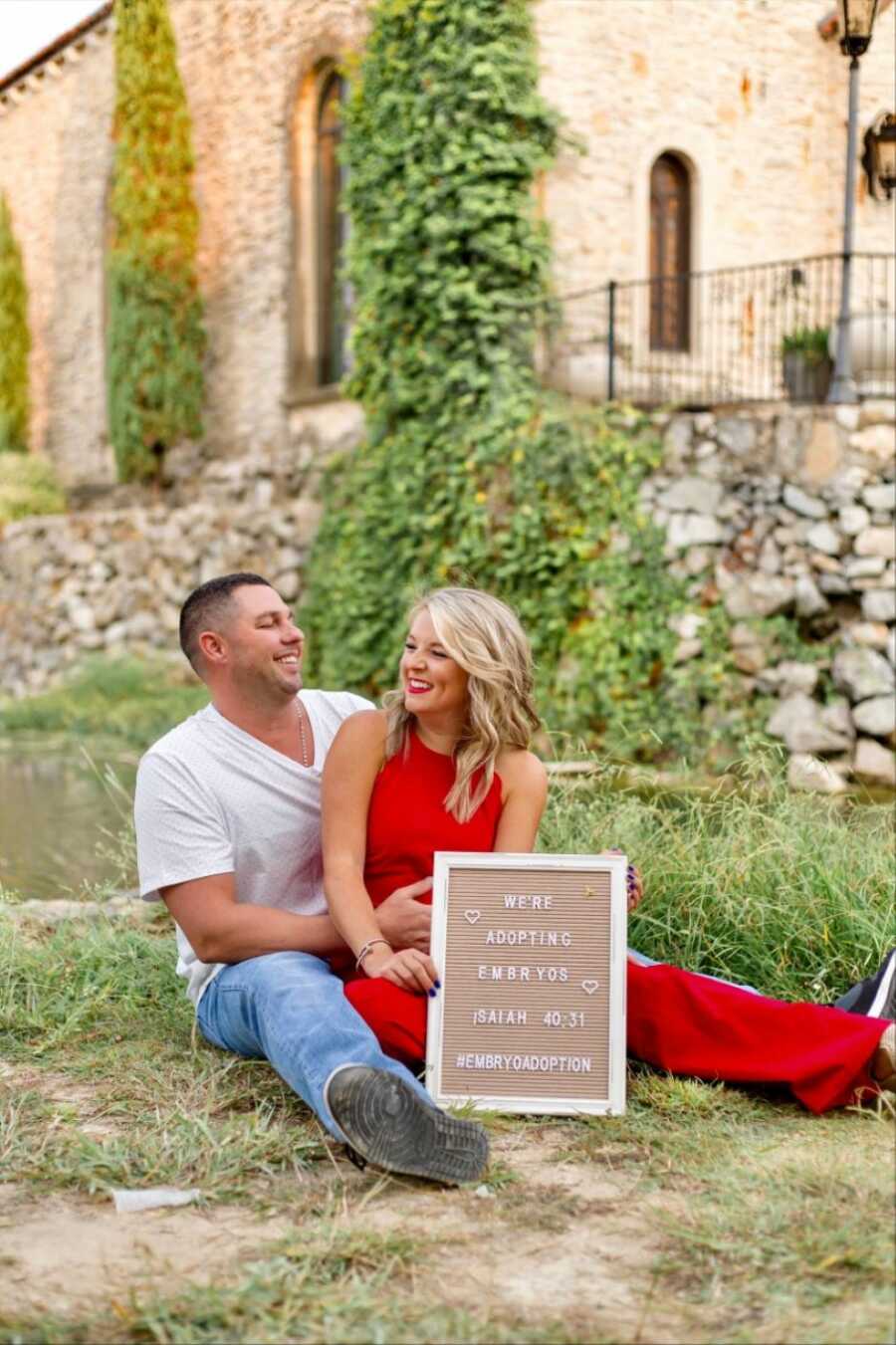 Young married couple smile at each other while sitting in the grass with a sign announcing that they're adopting embryos