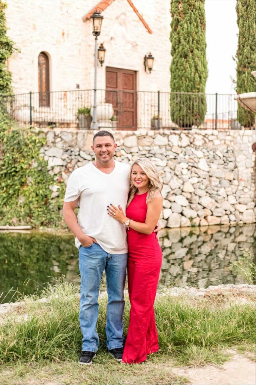 Young married couple smile big, the wife in a red romper and the husband in jeans and a white t-shirt, during an outdoor photoshoot