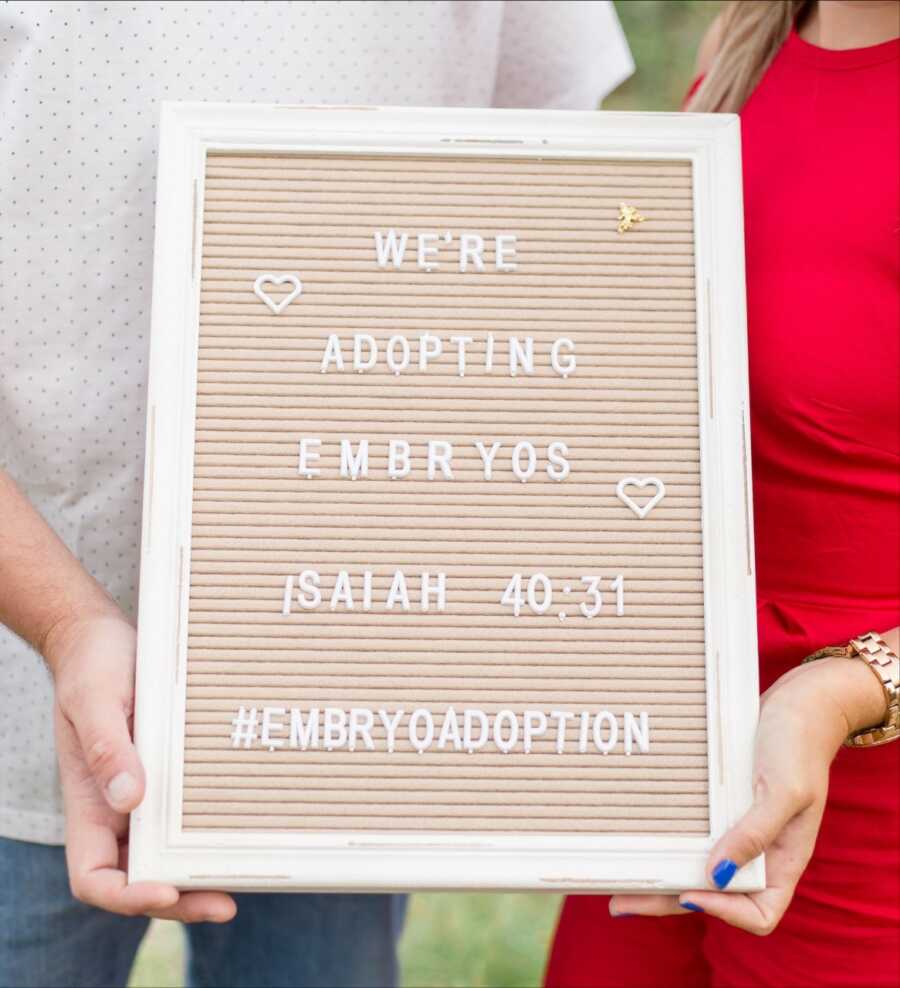 Married couple hold up a sign reading "We're adopting embryos" after struggling with infertility