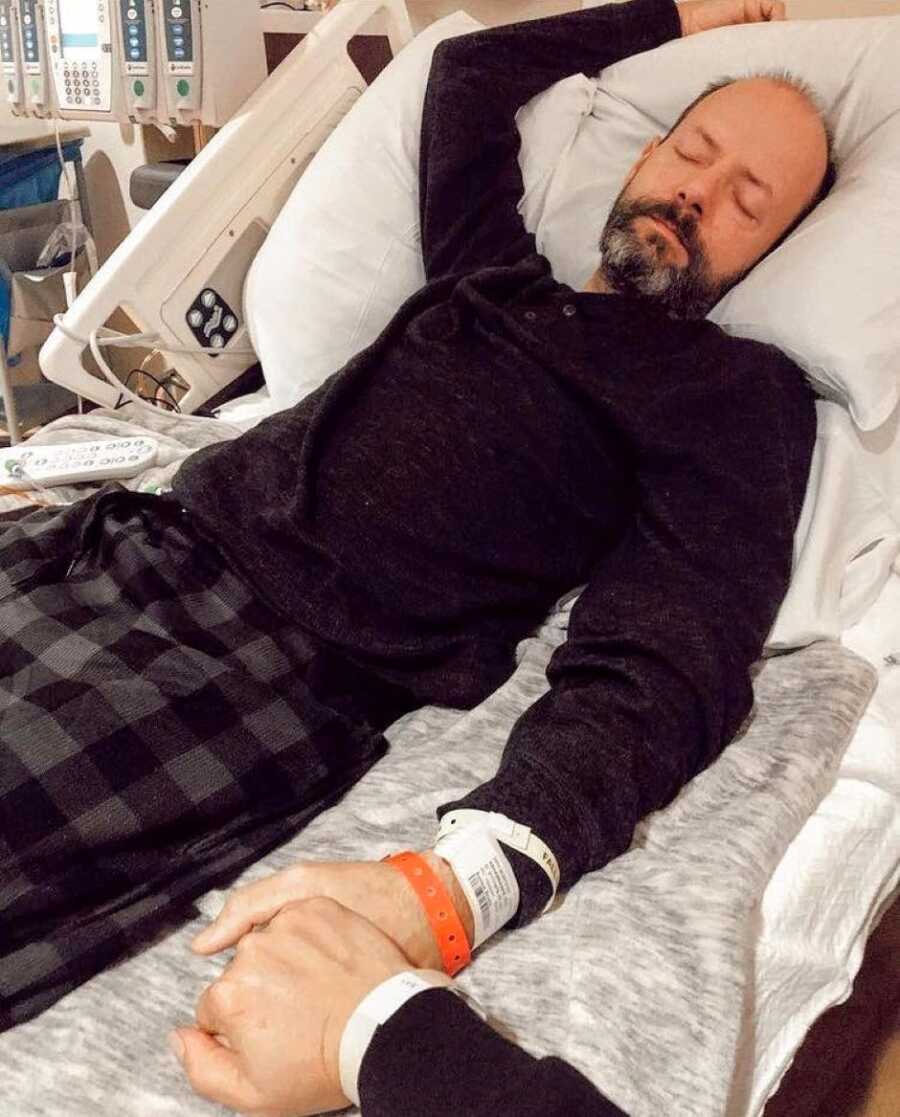 Woman holds hands with her husband dying of cancer while he lays in a hospital bed in black pajamas