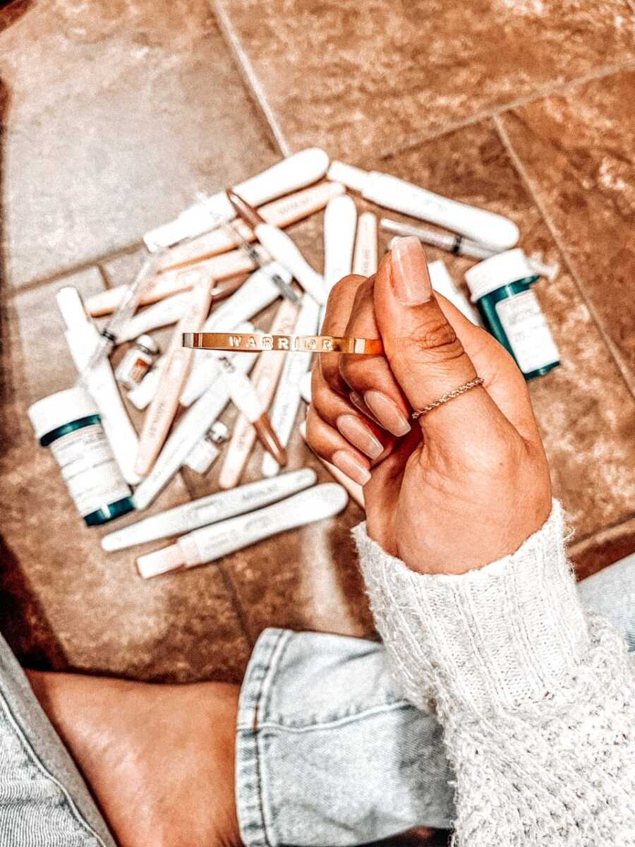 Woman trying to conceive takes a photo of a bracelet with "warrior" engraved in it above IVF needles, negative pregnancy tests, and pill bottles