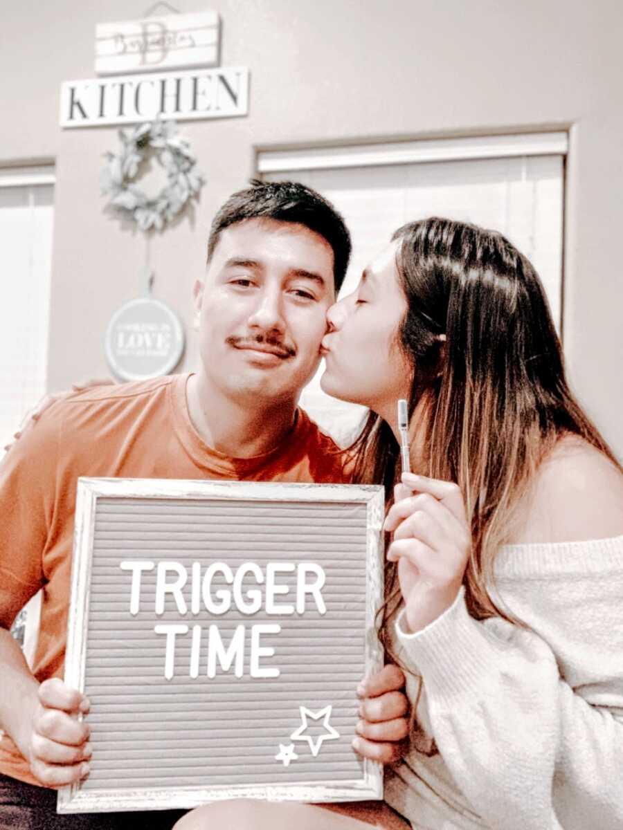 Woman kisses her partner's cheek while he holds a sign that says "trigger time" and she holds a needle for an IVF treatment