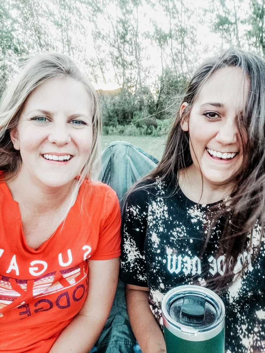 Mom and stepmom giggle as they share wine they snuck into their kids' football game in a Yeti cup