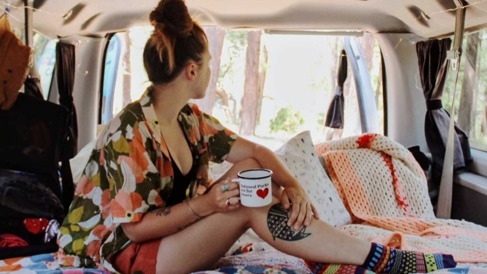 Van life traveler poses for an Instagram photo holding an empty coffee mug while sitting in the back of her van
