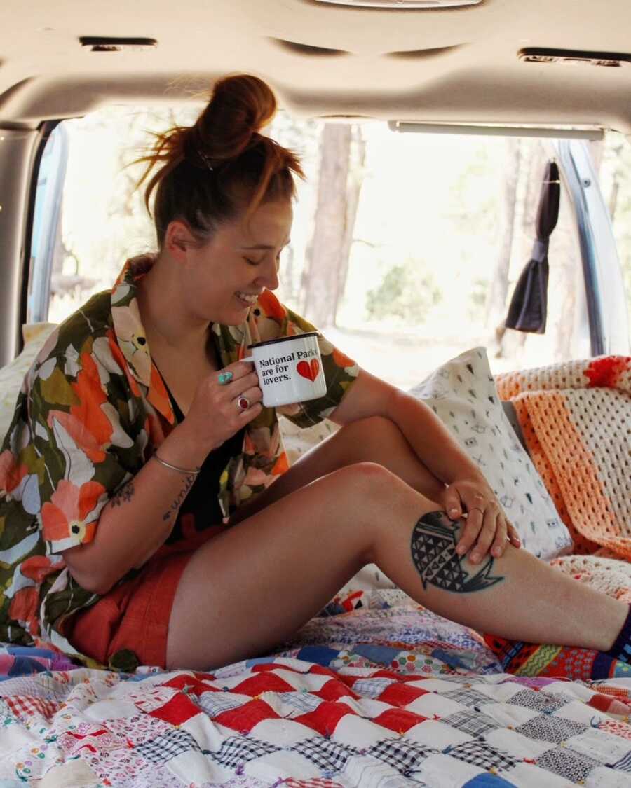 Van life traveler poses for a photo, fake laughing and holding an empty coffee cup, while sitting in the back of her van