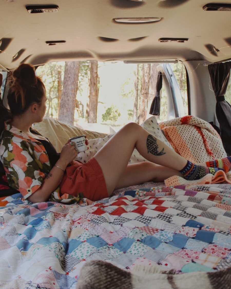 Van life traveler poses for a photo in the van she shares with her boyfriend while looking out at the trees