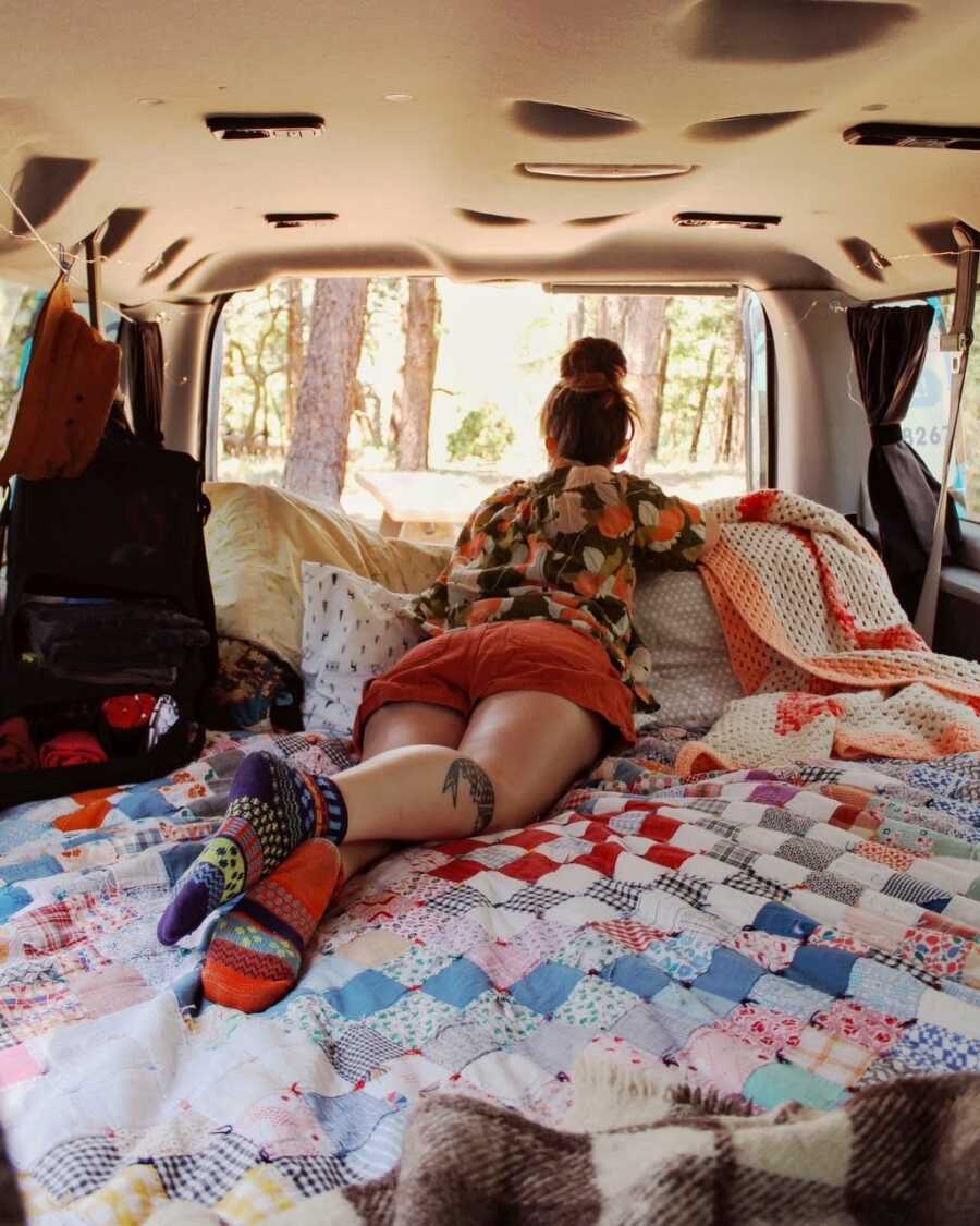 Woman lays on her stomach on a bunch of pillows while looking out of the back of her open van, taking in the view