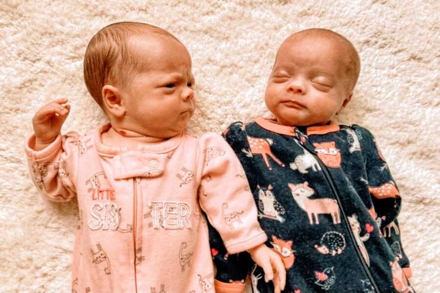 Newborn twin daughters wear fuzzy long-sleeved onesies while laying on a white fuzzy blanket, one asleep and one looking grumpy