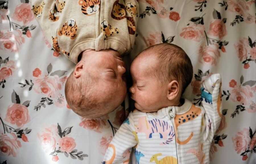 Twin girls wear onesies with different animals printed on them while napping on a floral blanket