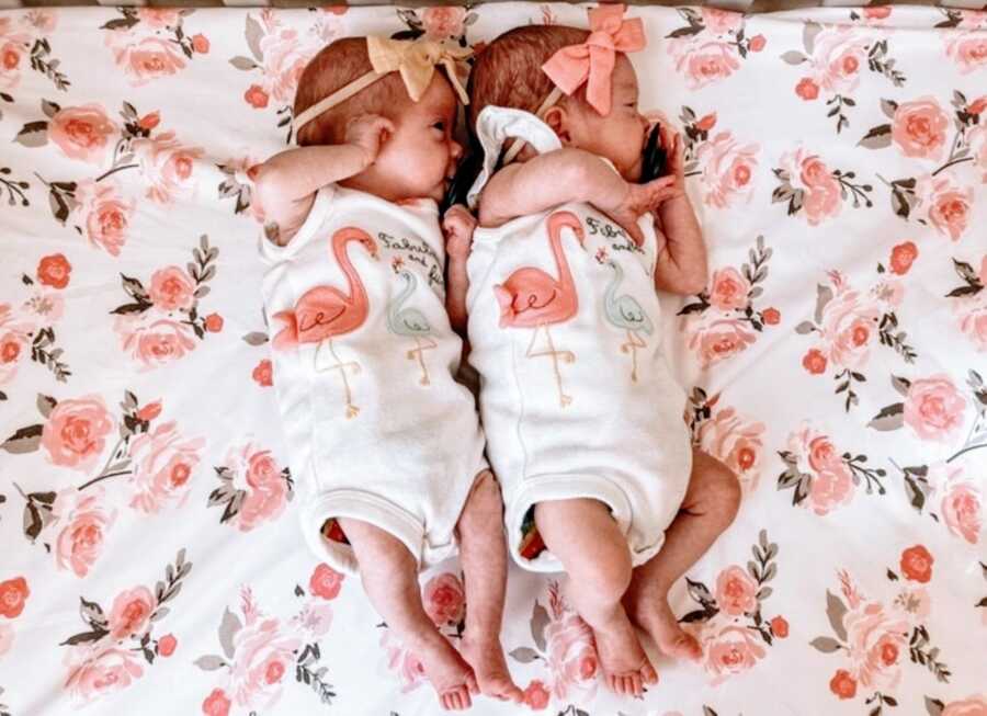 Twin newborn daughters wearing matching flamingo onesies with different colored bows on their heads