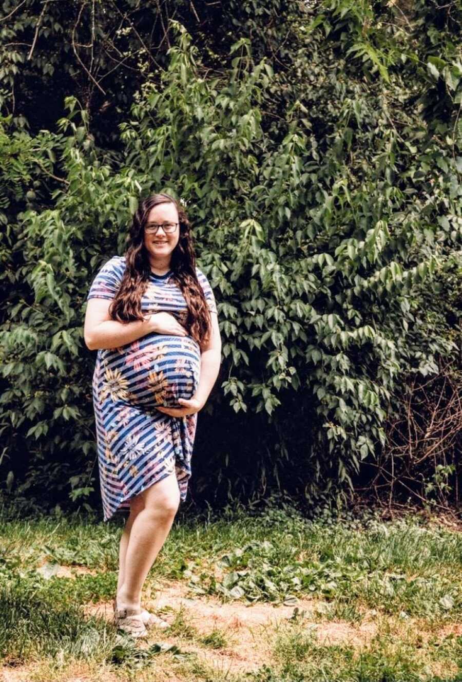 Woman pregnant with twin girls takes a photo of her belly bump in a blue-striped floral dress