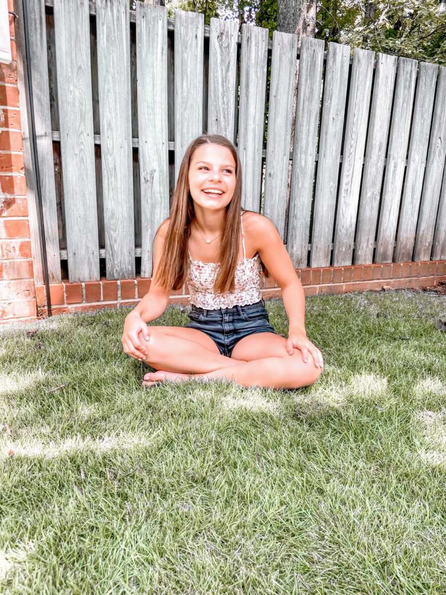 Young girl sits on the ground in her backyard and smiles while wearing a floral tank top and denim shorts