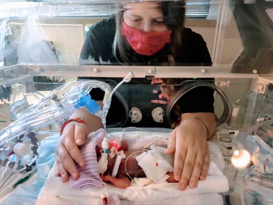 Mom wears a mask while touching her preemie child in the NICU while she's intubated in an isolette