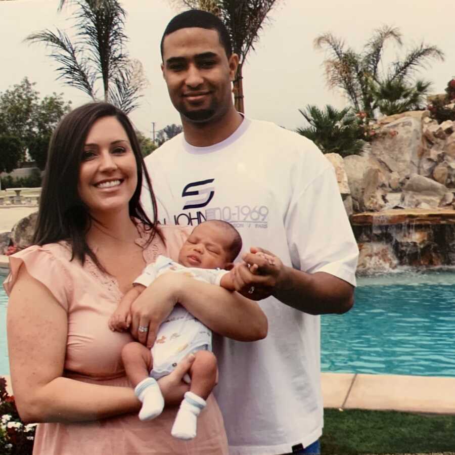 Young married couple and first time parents hold their newborn son outside with a pool behind them