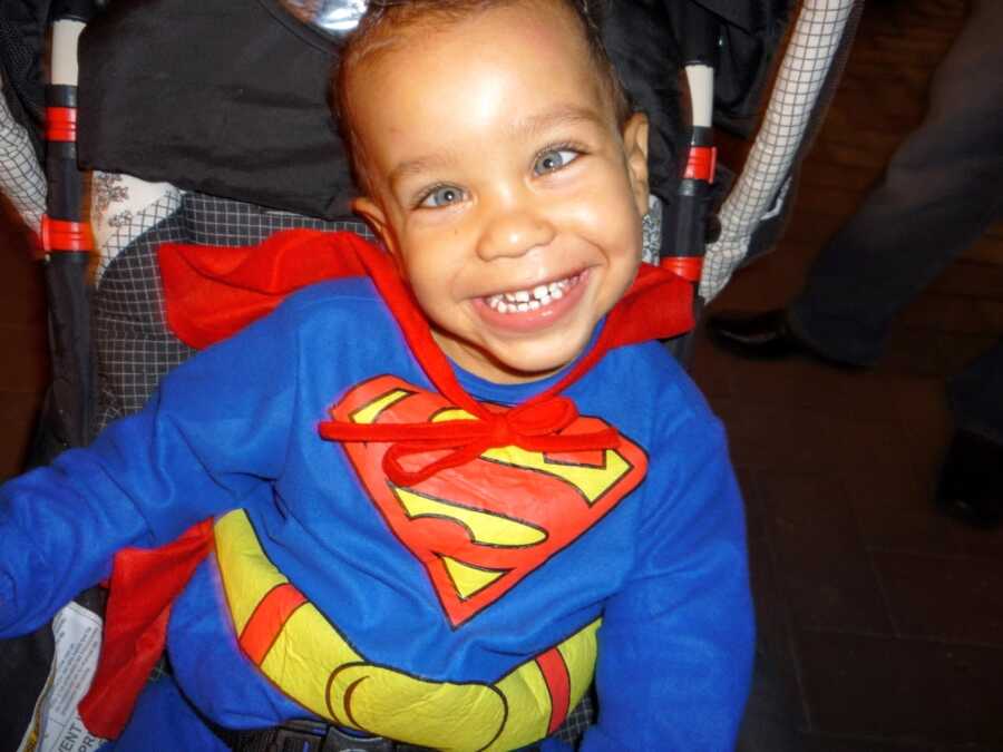 Little boy with Fragile X Syndrome smiles for a photo in his stroller while wearing a Superman suit