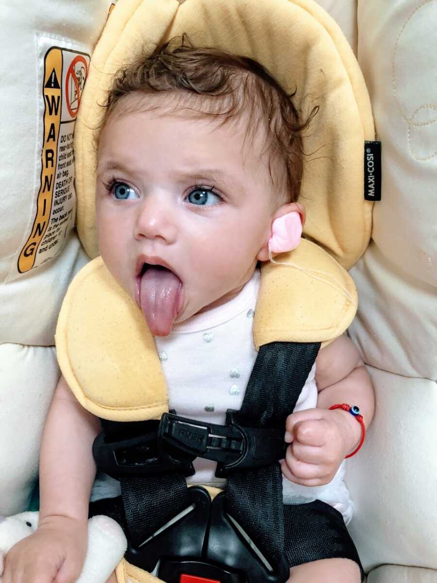 Little girl with blue eyes and hearing loss sticks out her tongue while sitting in a car seat with ear foam