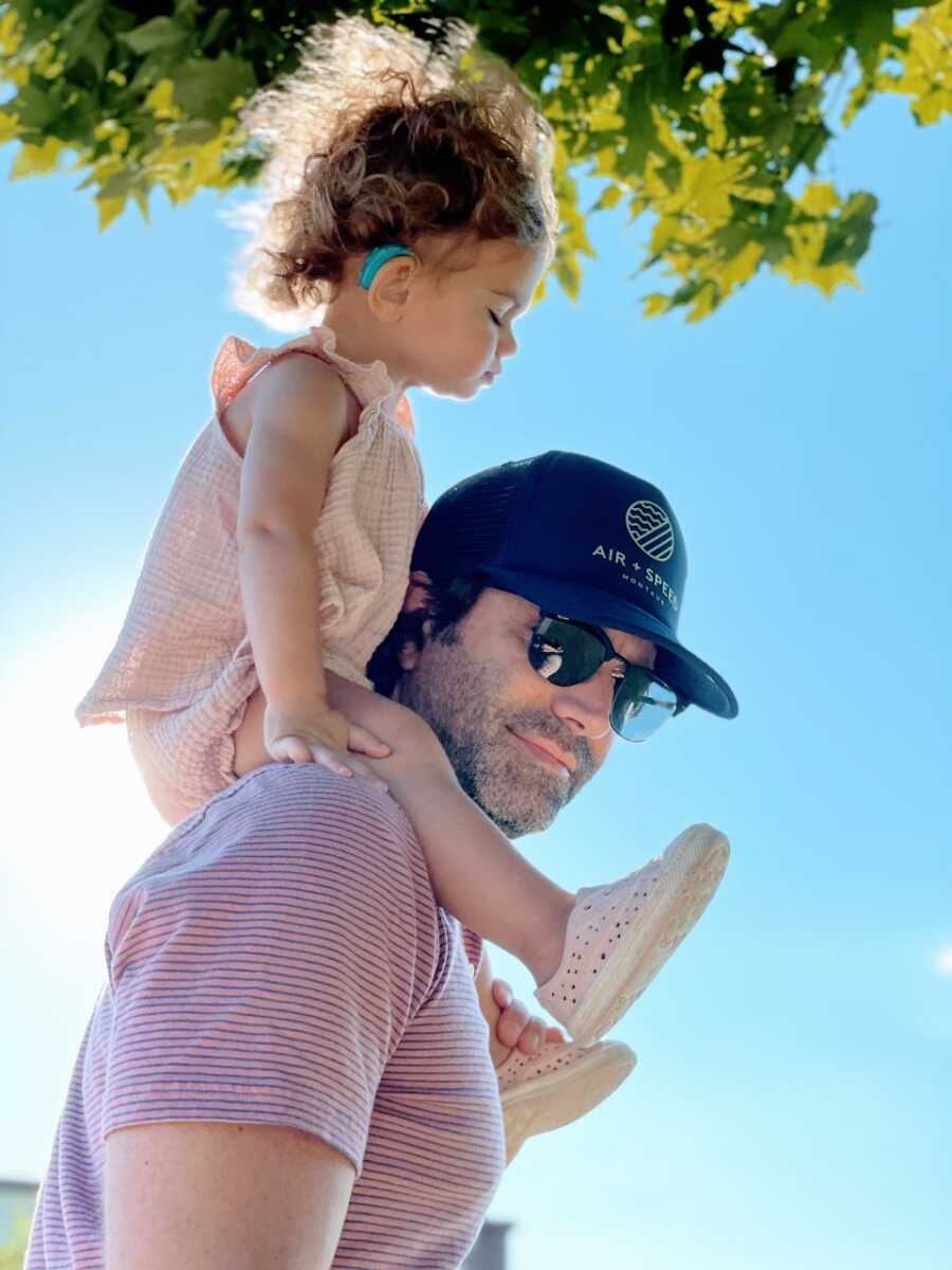 Dad carries his hard of hearing daughter on his shoulders, her blue cochlear implant visible