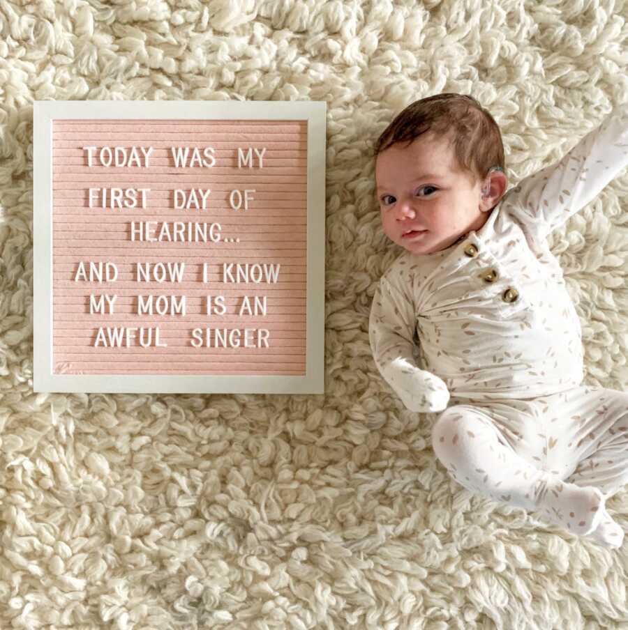 Young baby squirms around while wearing hearing aids next to a sign that reads "Today was my first day of hearing...and now I know my mom is an awful singer"