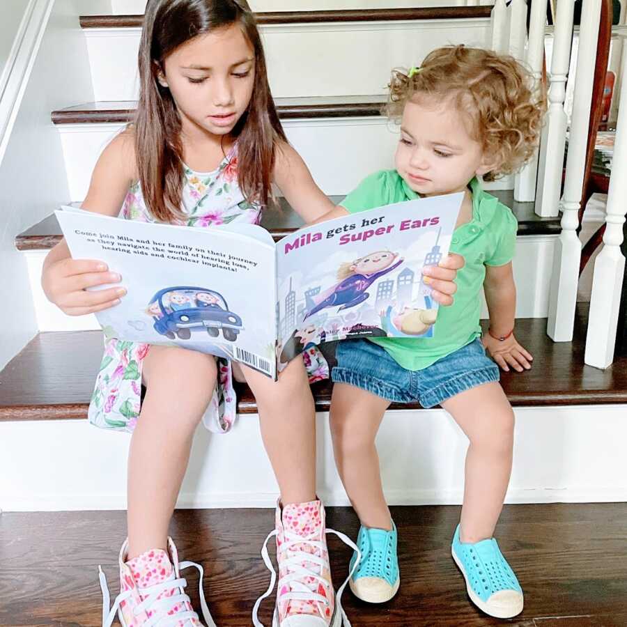 Big sister reads a book called "Mila Gets Her Super Ears" to her hard of hearing little sister