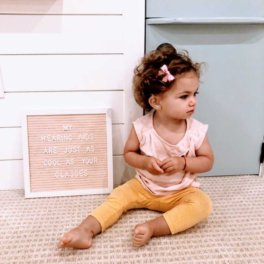 Little girl with hearing loss sits next a sign that reads "My hearing aids are just as cool as your glasses"