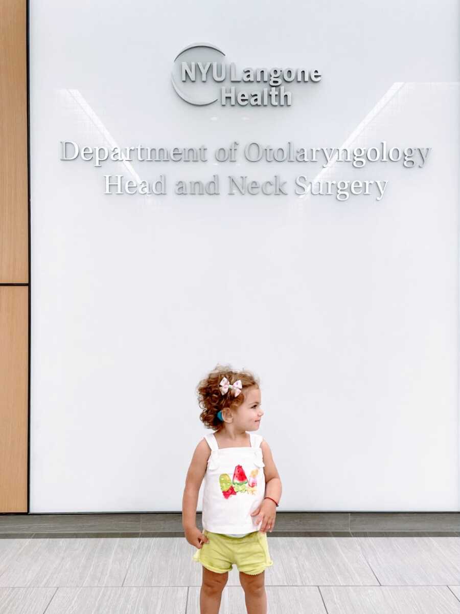 Little girl poses in front of a sign that reads "Department of Otolaryngology Head and Neck Surgery" with her new cochlear implants