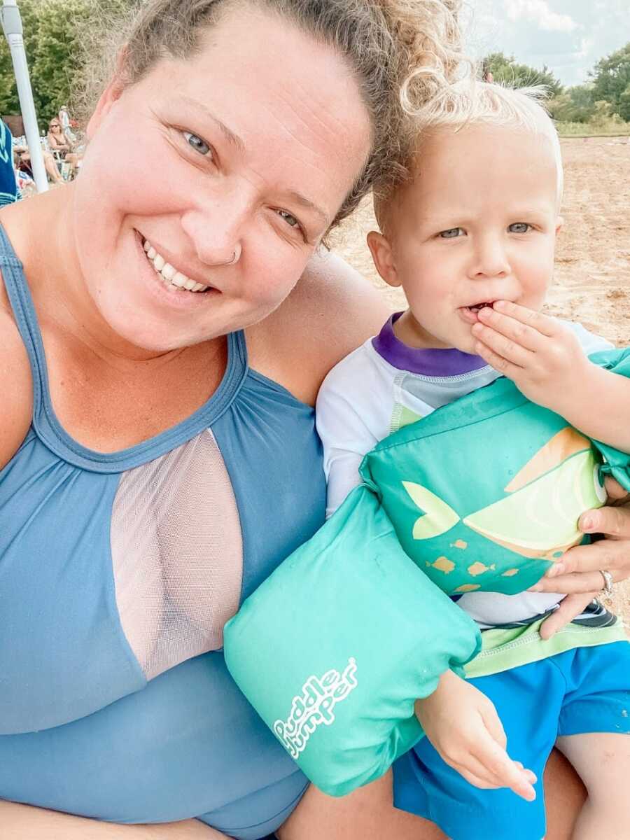 Mom of two smiles big in a blue one-piece bathing suit for a picture with her son while at the beach