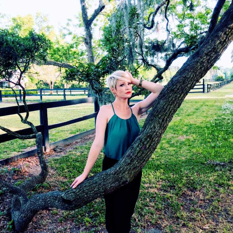 Working mom of three poses in front of a scaly bark tree in a green tank top, looking like a She-EO