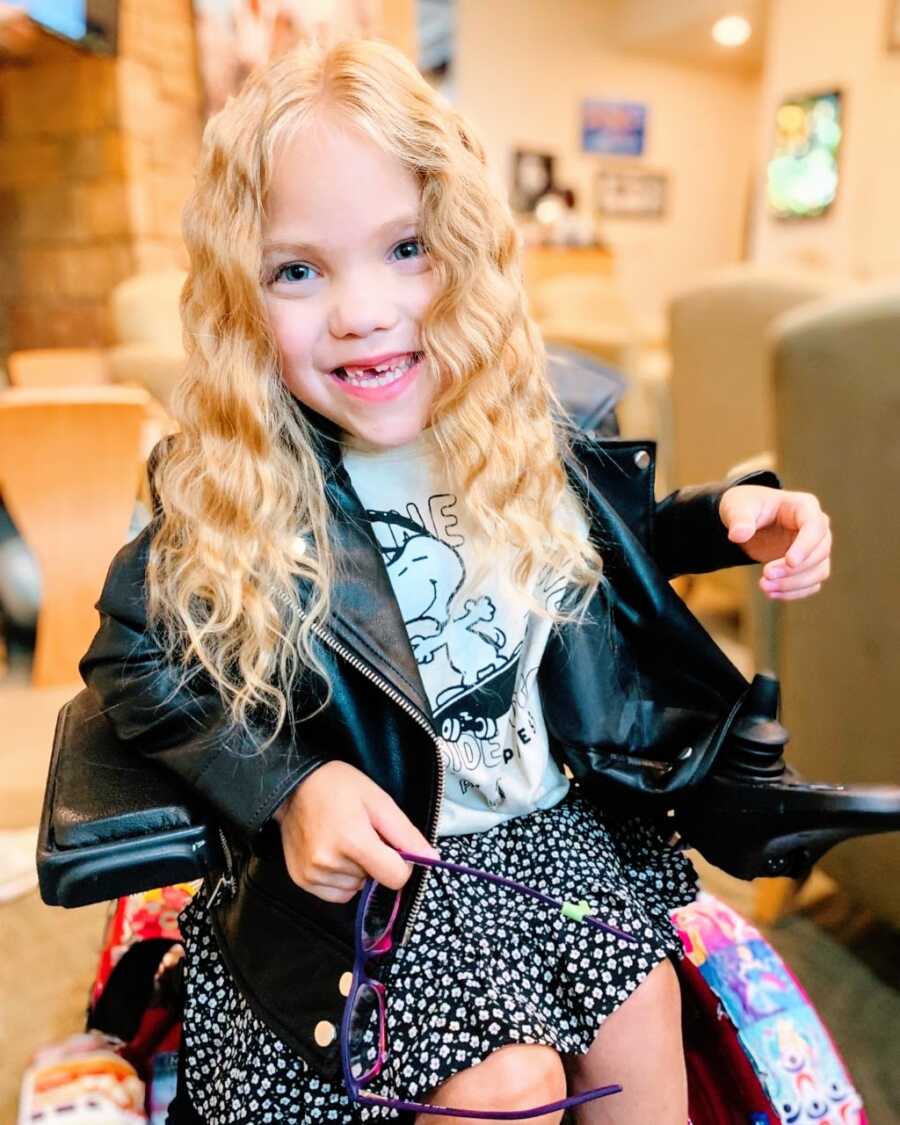 Little girl with cerebral palsy poses for a photo in her wheelchair with her hair curled while wearing a Snoopy shirt and a leather jacket