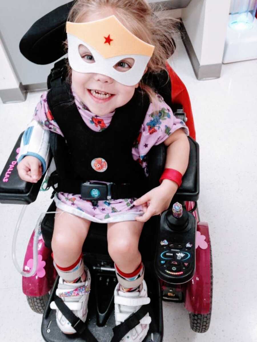 Little girl with cerebral palsy sits in her hot pink motorized wheelchair while wearing a superhero mask