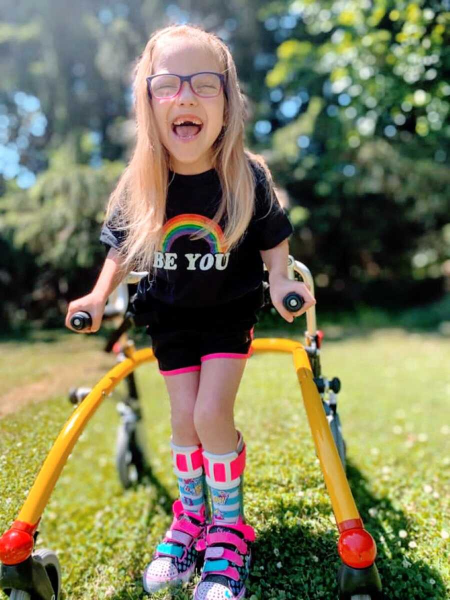 Little girl with cerebral palsy practices walking with a walker while wearing a shirt with a rainbow on it that says "Be You"