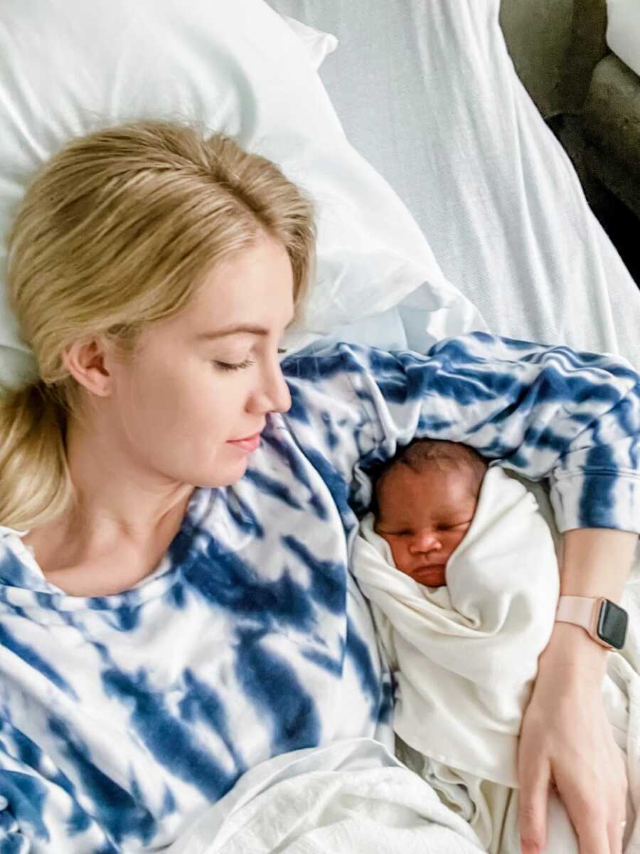 Woman wearing a blue and white tie-dyed shirt looks down at her newborn adopted son while they lay in bed together